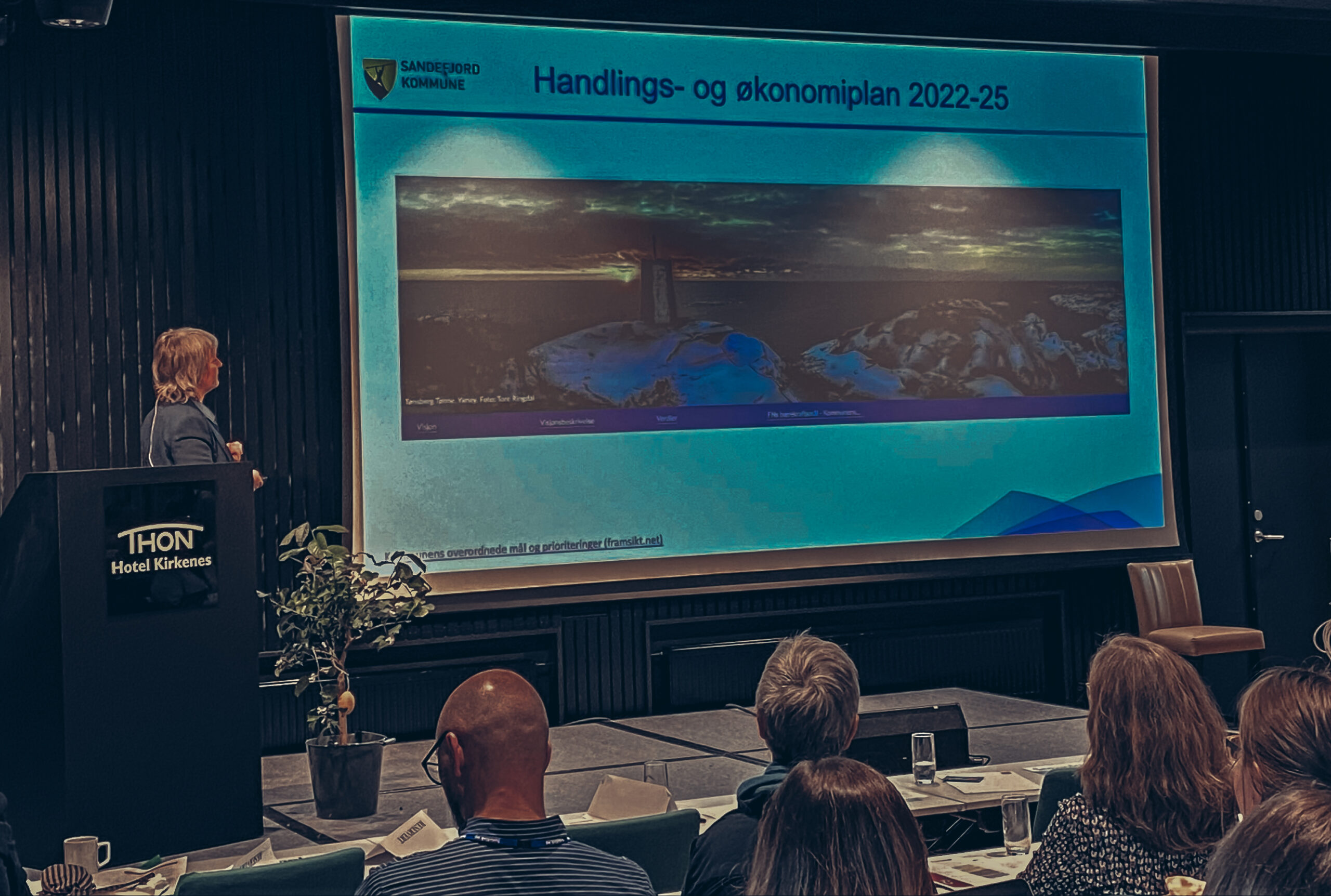 Anne Therese Anvik, fra Sandefjord kommune, står på scenene under Kommunalplankonferanse 2022. På skjerm vies en powerpoint med bilde av deres handlings- og økonomiplanen, utarbeidet i Framsikt. 