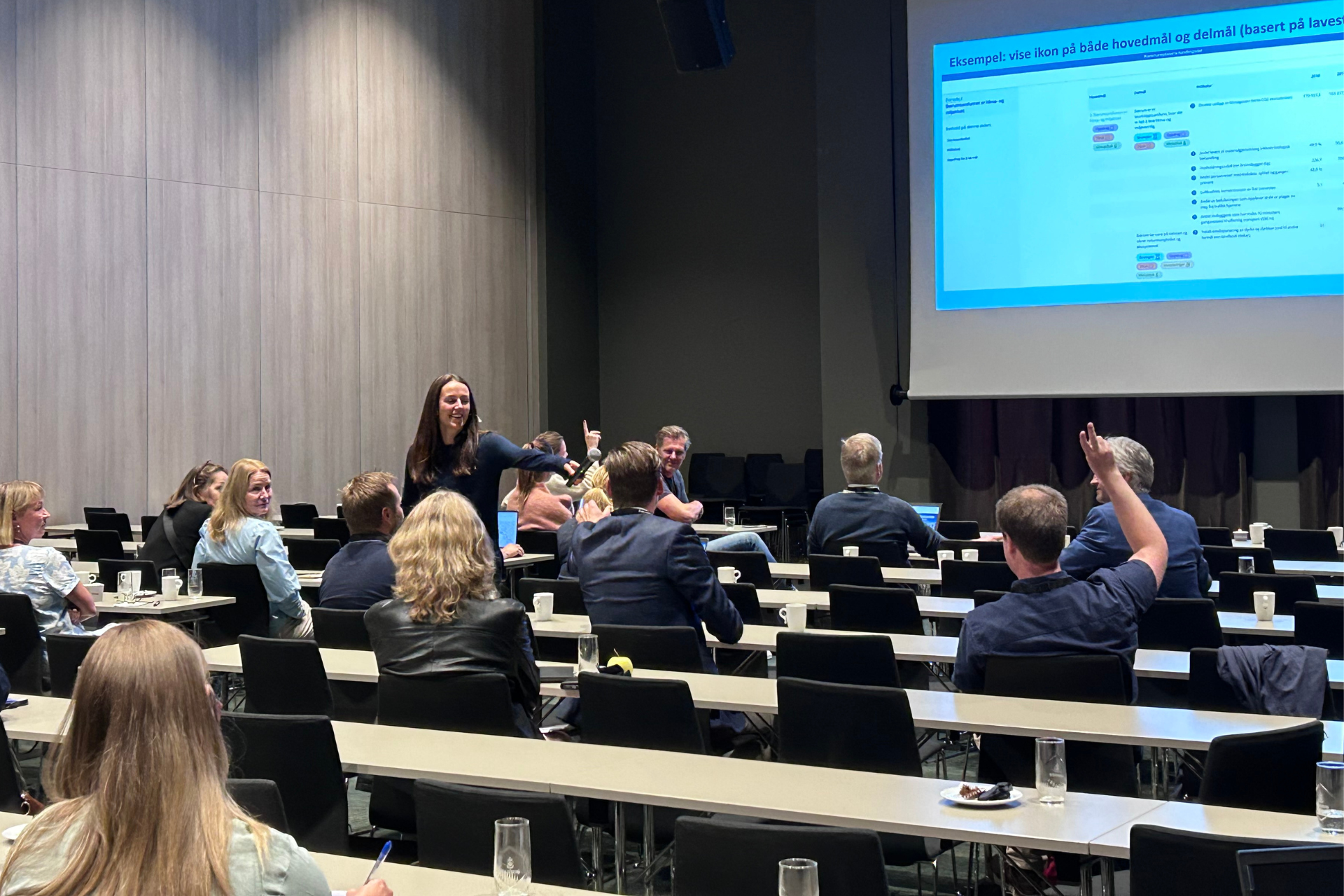 En dame går rundt med mikrofon i en forsamling. En mann rekker opp hånden. 
