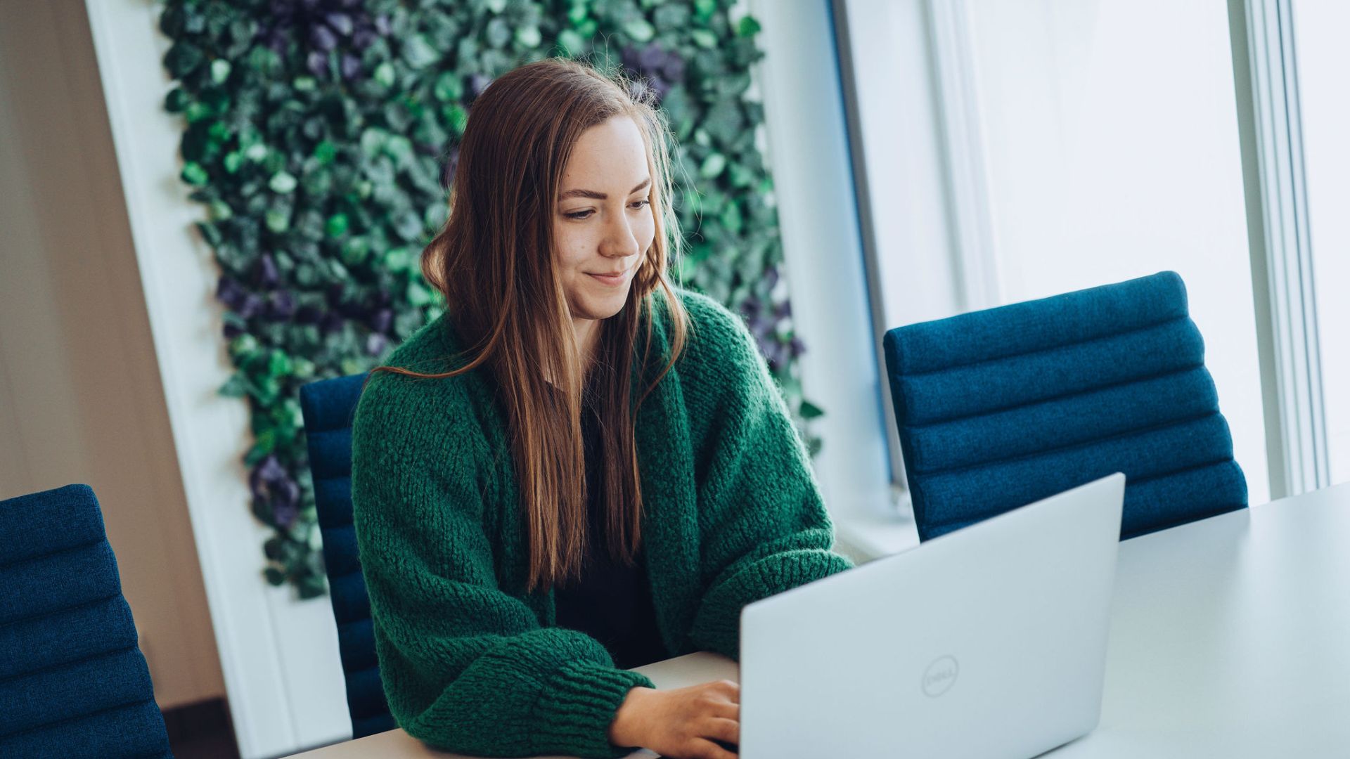 En kvinne med langt brunt hår som sitter ved et bord med en bærbar datamaskin. Hun er iført en grønn cardigan og ser fokusert ut mens hun jobber. I bakgrunnen er det en veggdekorasjon med grønt og lilla bladverk, og rommet er lyst med naturlig lys som strømmer inn fra store vinduer.