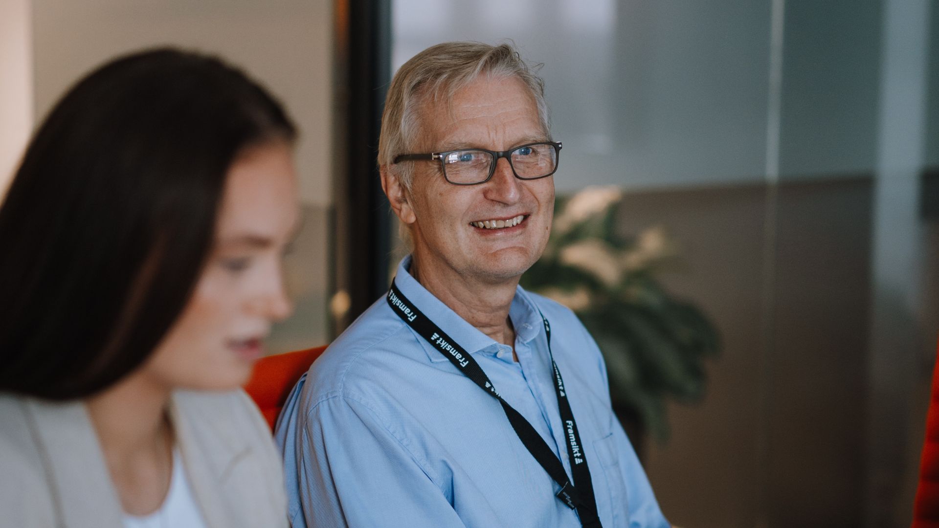 En mann med grått hår som smiler mens han sitter ved et bord i et møtelignende miljø. Han har på seg en blå skjorte og briller, med en ID-brikke hengende rundt halsen. Ved siden av ham er en kvinne med langt brunt hår, uskarp i forkant av bildet. 