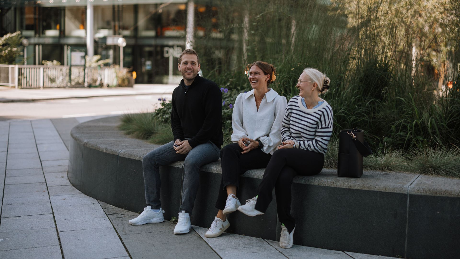 Tre voksne smiler og sitter på en steinmur utendørs. En mann til venstre, en kvinne i midten, og en kvinne til høyre. Bak dem er det grønne planter og bygninger.