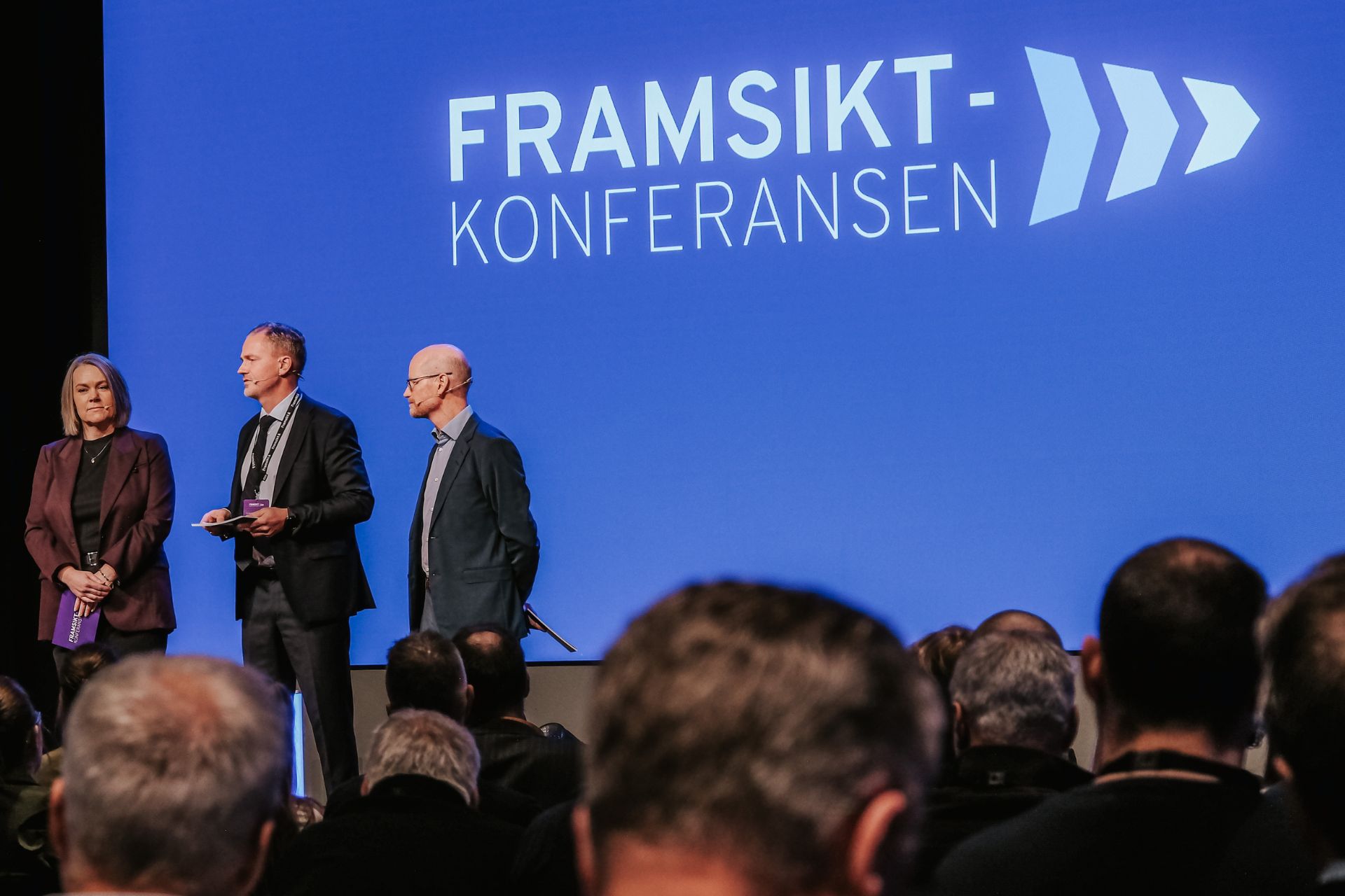 3 personer som står på scenen og holder Framsiktkonferansen med masse folk i publikum.
