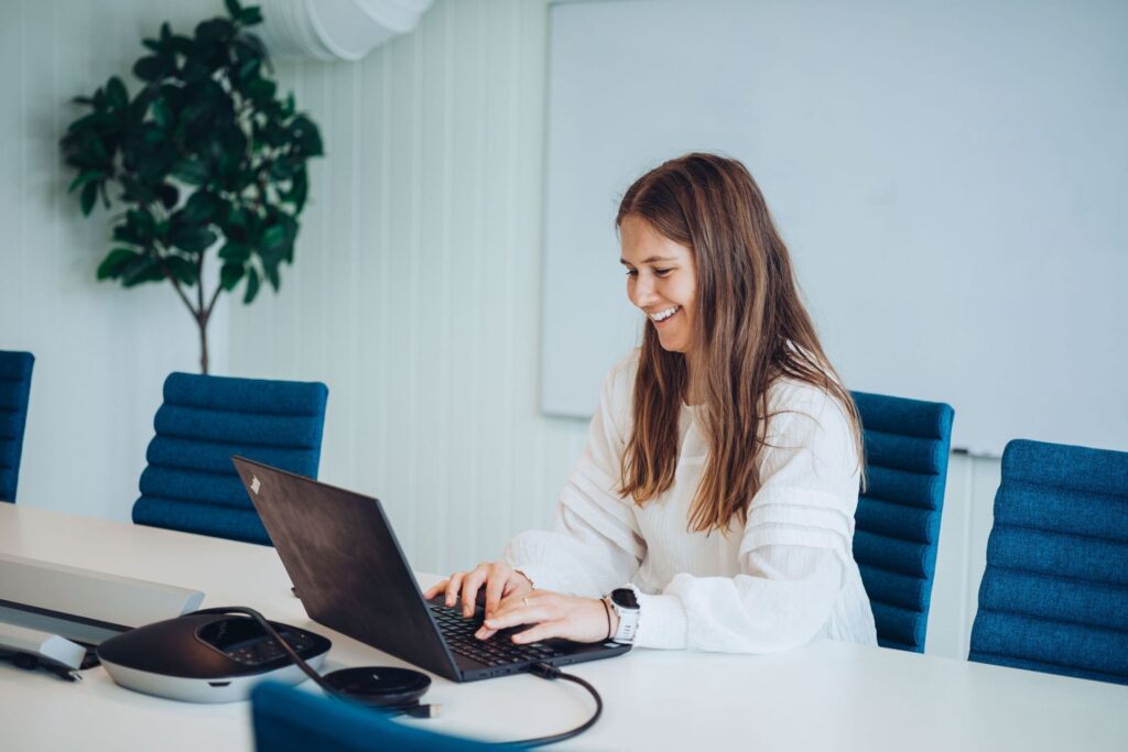 En dame som sitter på en stol foran en bærbar PC på et bord og skriver med tastaturet. Hun ser på skjermen og smiler.